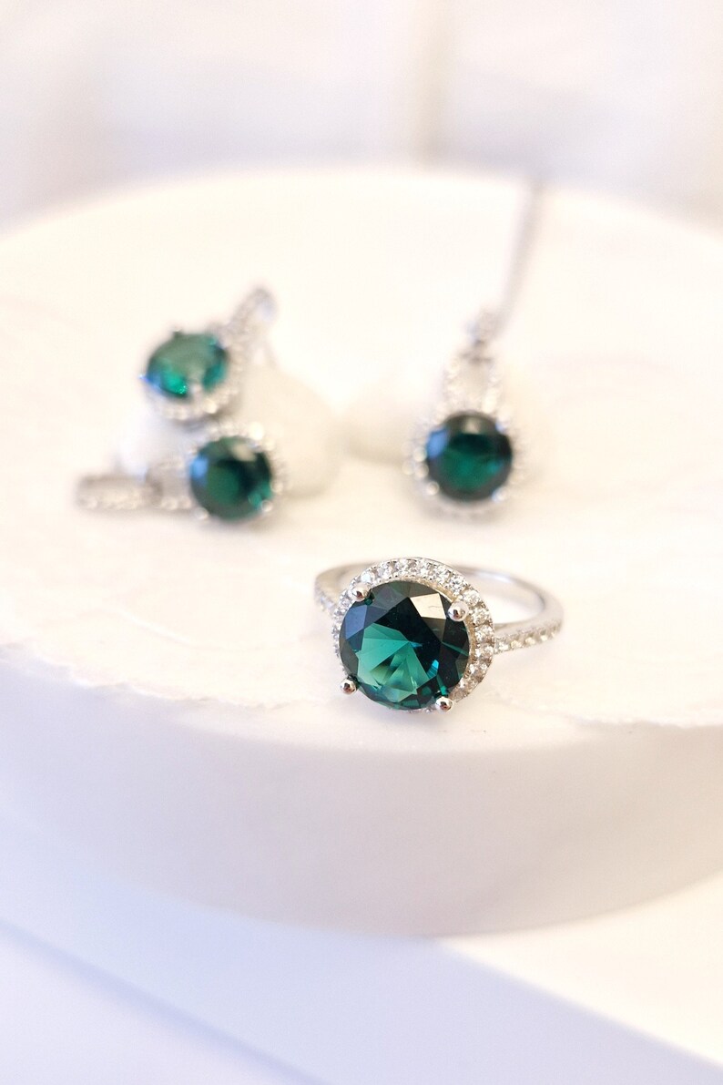 May include: A silver ring with a large, round, dark green gemstone surrounded by a halo of smaller, clear gemstones.