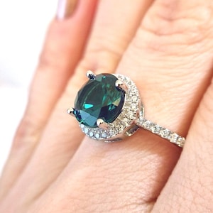 May include: A silver ring with a large, round, dark green gemstone surrounded by a halo of smaller, clear gemstones. The ring has a thin band with smaller clear gemstones.