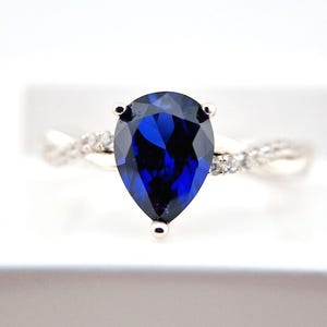 May include: A silver ring with a pear-shaped blue sapphire gemstone and smaller clear gemstones on the band.