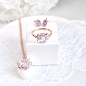 Peut inclure: Un ensemble de bijoux en or rose comprenant une bague, des boucles d'oreilles et un collier. Chaque pi&egrave;ce est orn&eacute;e d'une pierre hexagonale violet clair et de petites pierres claires. L'ensemble est pr&eacute;sent&eacute; sur une surface blanche.