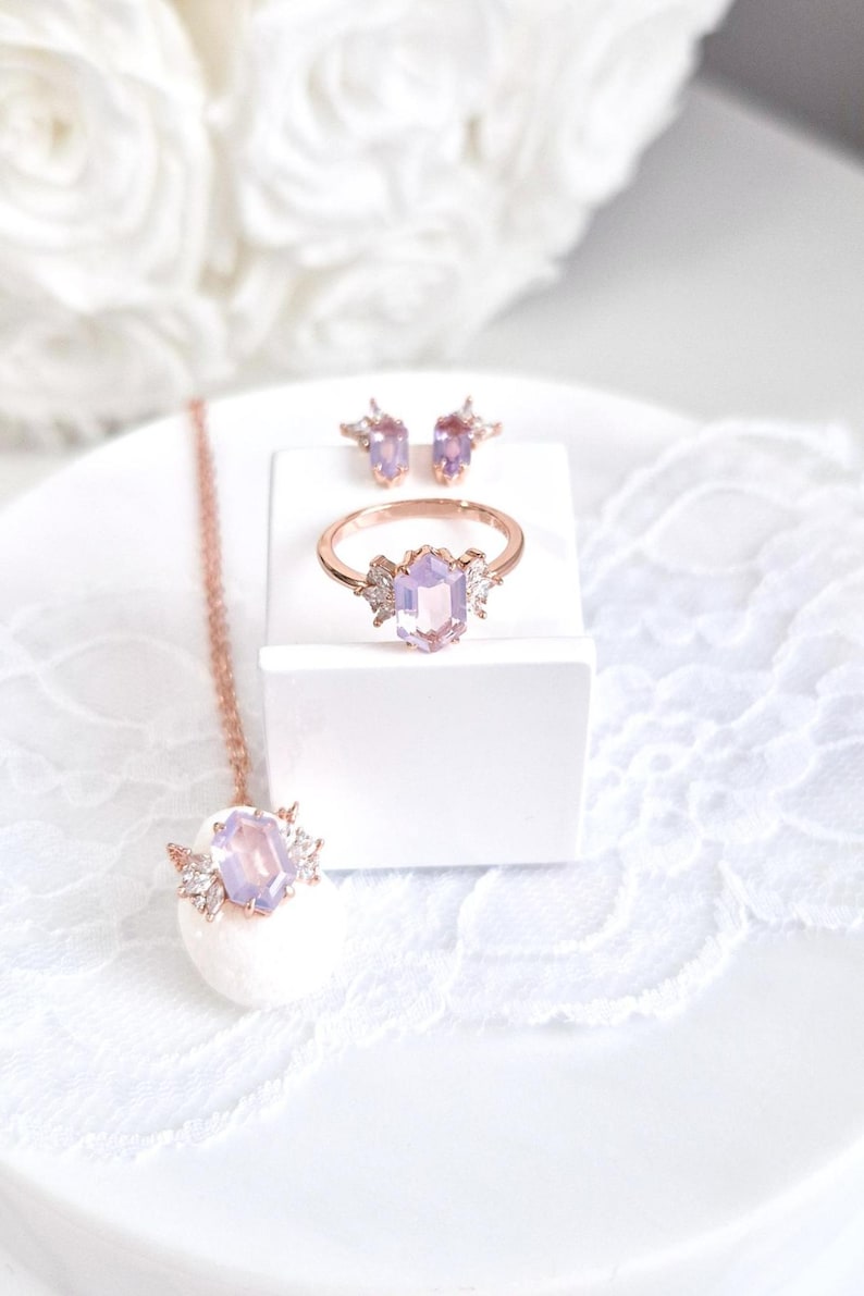 Peut inclure: Un ensemble de bijoux en or rose comprenant une bague, des boucles d'oreilles et un collier. Chaque pi&egrave;ce est orn&eacute;e d'une pierre hexagonale violet clair et de petites pierres claires. L'ensemble est pr&eacute;sent&eacute; sur une surface blanche.