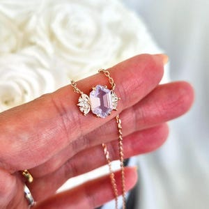May include: A delicate gold necklace featuring a large, light purple gemstone with an emerald cut. The gemstone is flanked by smaller, clear crystals. The necklace is held up by a hand.