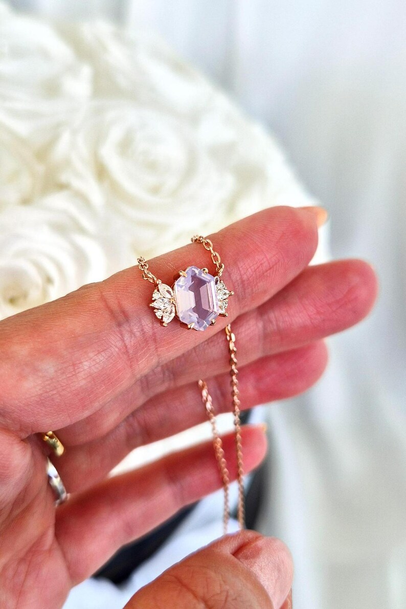 May include: A delicate gold necklace featuring a large, light purple gemstone with an emerald cut. The gemstone is flanked by smaller, clear crystals. The necklace is held up by a hand.