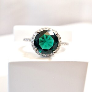 May include: A silver ring with a large, round, dark green gemstone surrounded by a halo of smaller, clear gemstones.