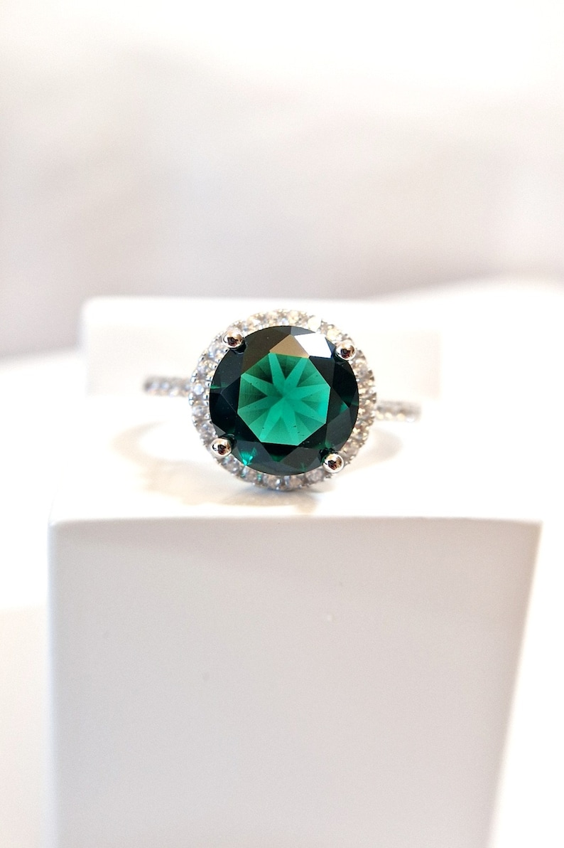 May include: A silver ring with a large, round, dark green gemstone surrounded by a halo of smaller, clear gemstones.