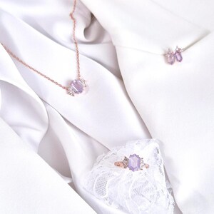 Peut inclure: Ensemble de bijoux en or rose comprenant un collier, une bague et des boucles d'oreilles, chacun orn&eacute; d'une grosse pierre pr&eacute;cieuse violet clair et de petites pierres transparentes. Les bijoux sont pr&eacute;sent&eacute;s sur un tissu blanc et soyeux.