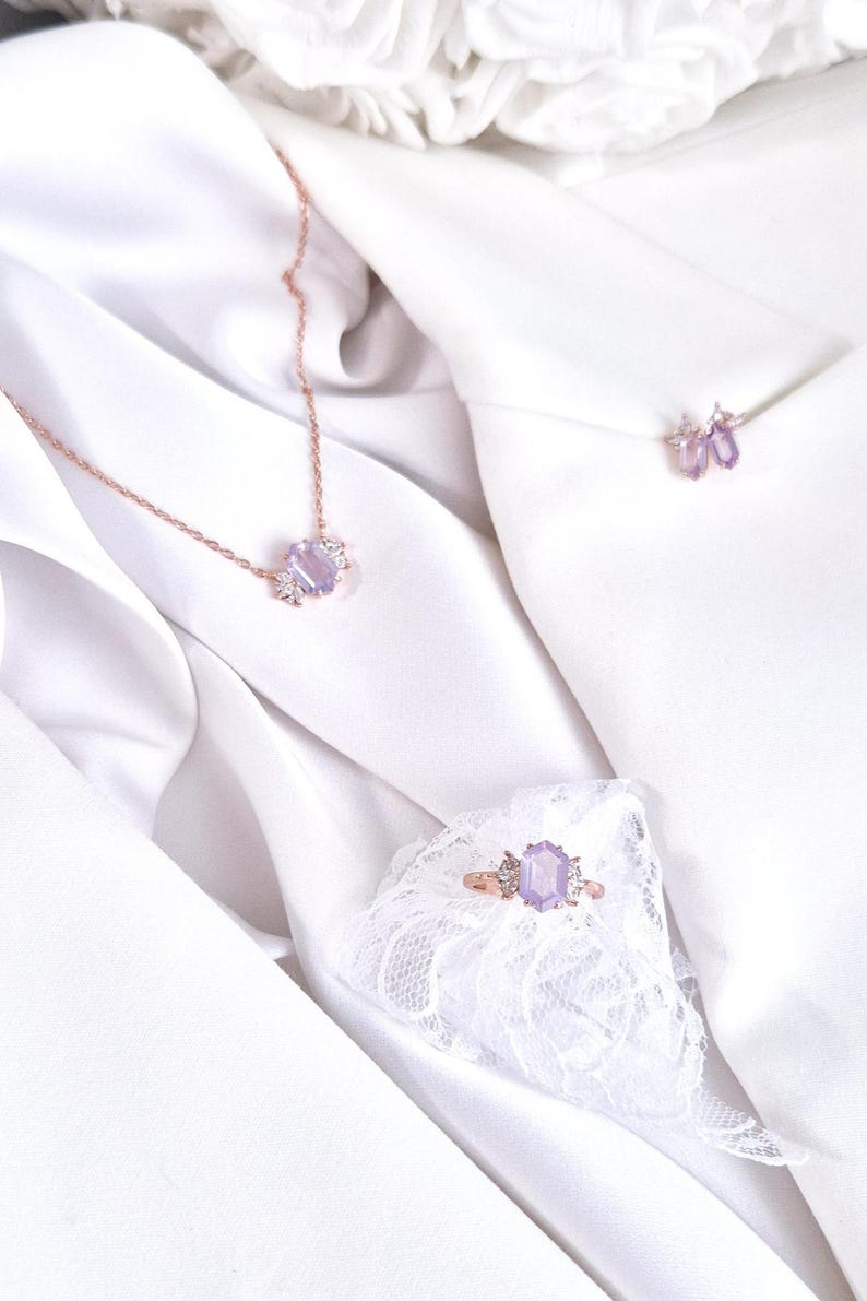 Peut inclure: Ensemble de bijoux en or rose comprenant un collier, une bague et des boucles d'oreilles, chacun orn&eacute; d'une grosse pierre pr&eacute;cieuse violet clair et de petites pierres transparentes. Les bijoux sont pr&eacute;sent&eacute;s sur un tissu blanc et soyeux.