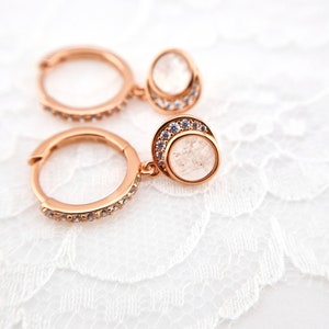 May include: Rose gold hoop earrings with a dangling clear stone set in a rose gold bezel with a row of clear stones around the edge.