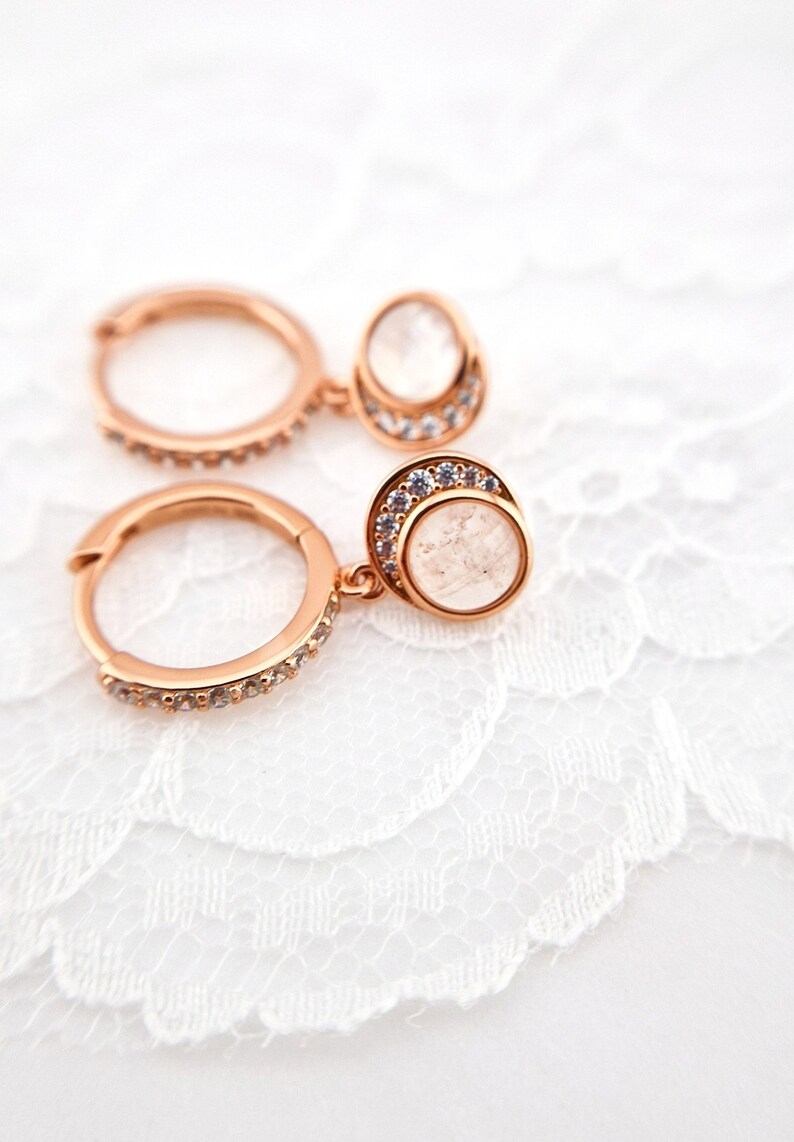 May include: Rose gold hoop earrings with a dangling clear stone set in a rose gold bezel with a row of clear stones around the edge.