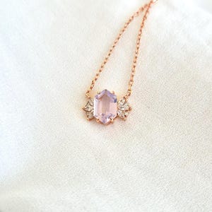 May include: A rose gold necklace featuring a hexagonal, light purple gemstone pendant. The pendant is flanked by two decorative, clear crystal accents. The necklace chain is delicate and rests on a white, textured surface.