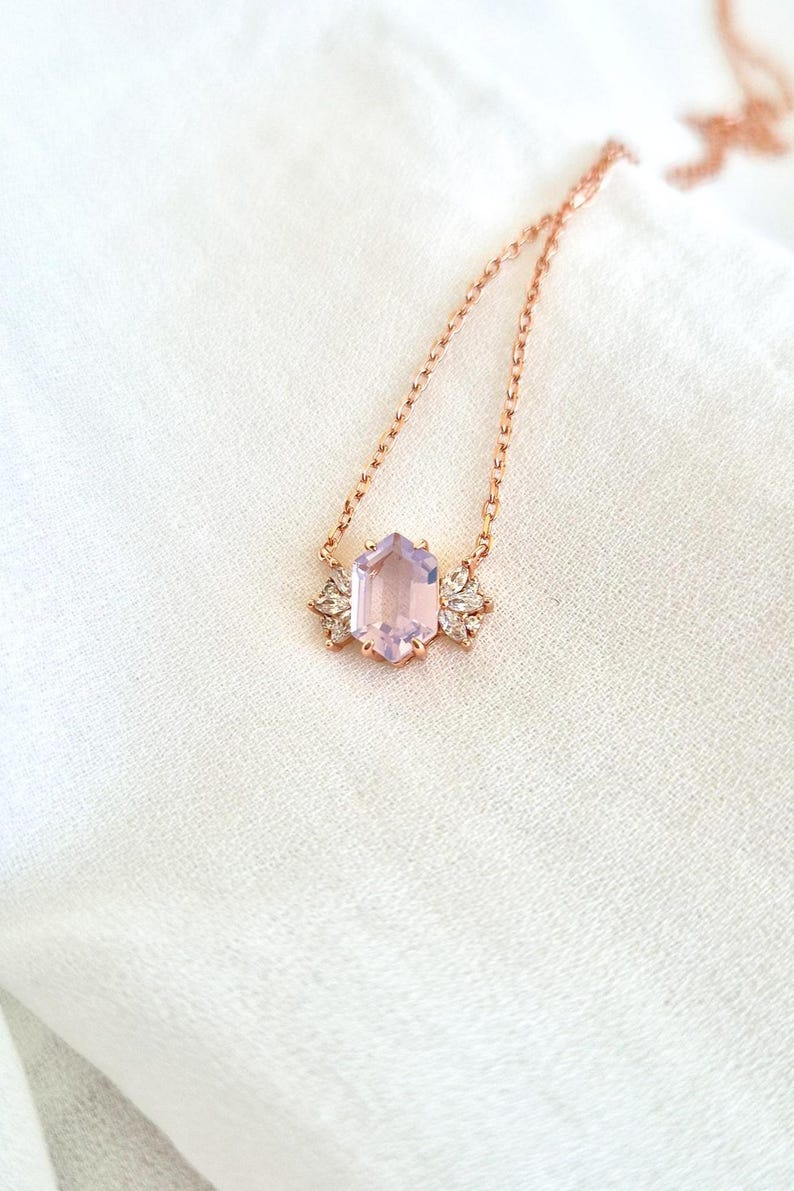 May include: A rose gold necklace featuring a hexagonal, light purple gemstone pendant. The pendant is flanked by two decorative, clear crystal accents. The necklace chain is delicate and rests on a white, textured surface.