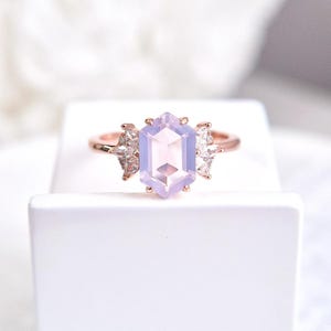 Peut inclure: Une bague en or rose orn&eacute;e d'une grande pierre hexagonale violet clair en son centre. La bague est flanqu&eacute;e de petites pierres claires en forme de poire de chaque c&ocirc;t&eacute; de la pierre principale. La bague est pos&eacute;e sur une surface blanche.
