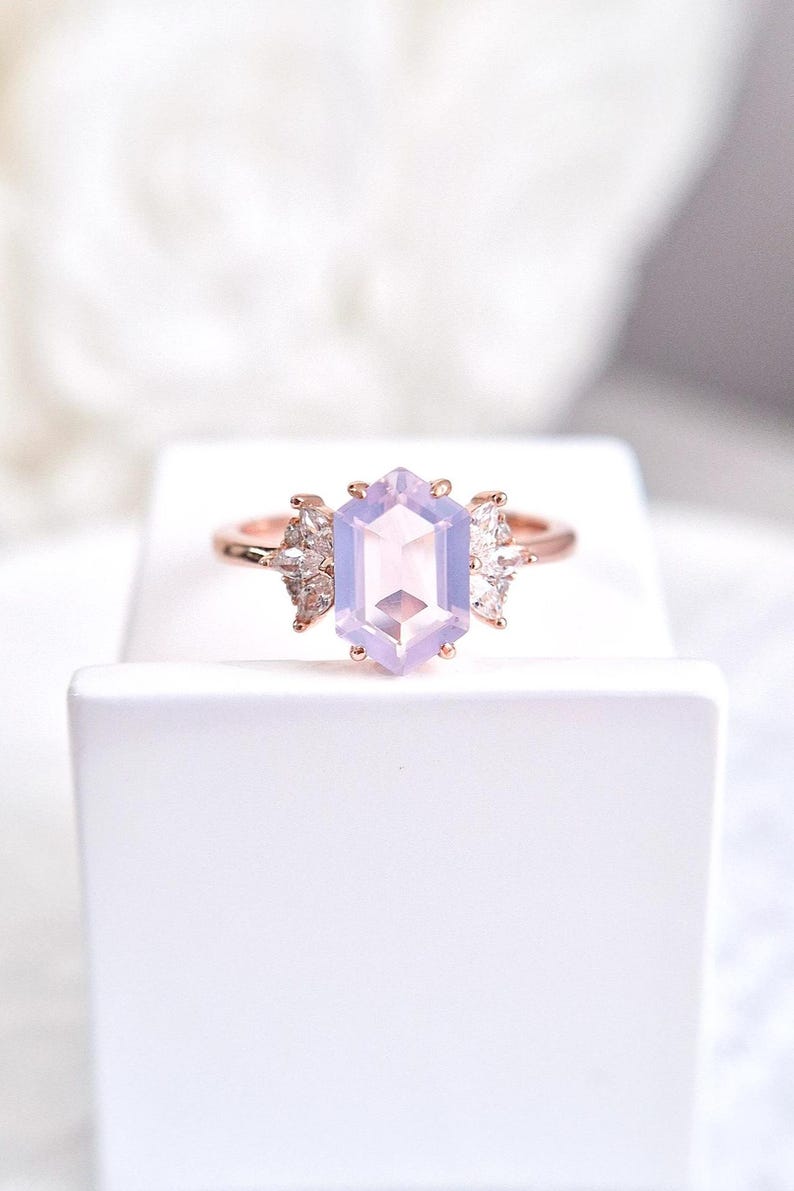 Peut inclure: Une bague en or rose orn&eacute;e d'une grande pierre hexagonale violet clair en son centre. La bague est flanqu&eacute;e de petites pierres claires en forme de poire de chaque c&ocirc;t&eacute; de la pierre principale. La bague est pos&eacute;e sur une surface blanche.
