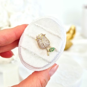 May include: A gold-tone tulip brooch with clear and green cubic zirconia stones. The brooch is displayed on a white velvet cushion.