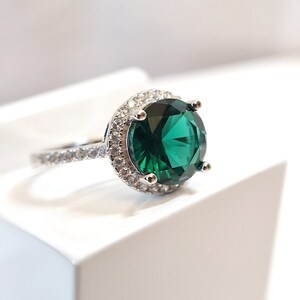 May include: A silver ring with a large, round, emerald green gemstone surrounded by a halo of smaller, clear gemstones.
