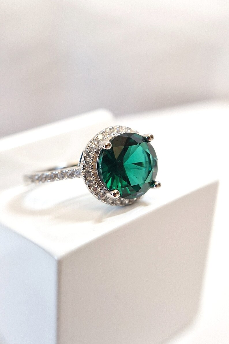 May include: A silver ring with a large, round, emerald green gemstone surrounded by a halo of smaller, clear gemstones.