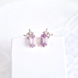 Peut inclure: Boucles d'oreilles en or rose avec des pierres rectangulaires violet clair. Chaque boucle d'oreille comporte un groupe de petits cristaux transparents au-dessus de la pierre principale. Les boucles d'oreilles sont pr&eacute;sent&eacute;es sur une surface blanche.
