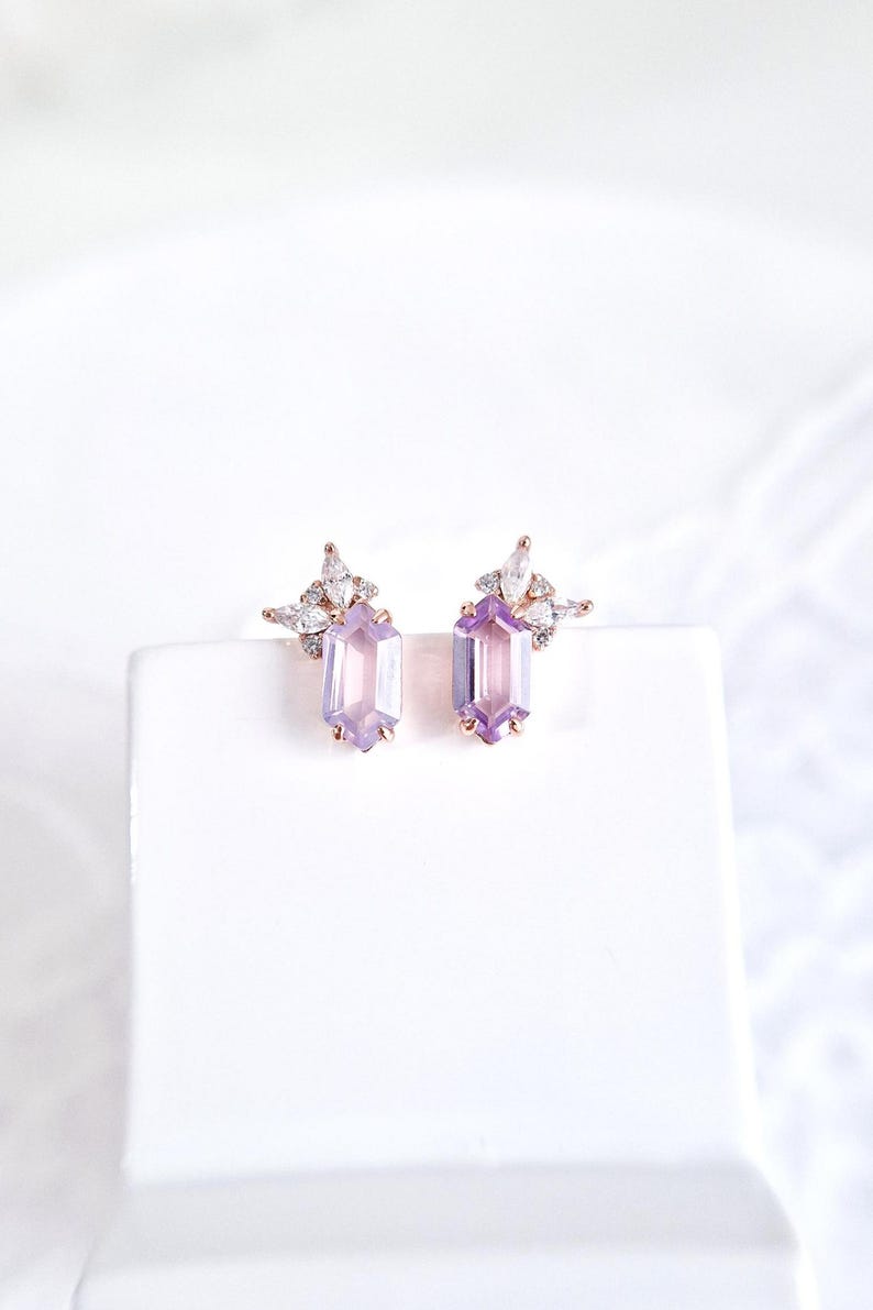 Peut inclure: Boucles d'oreilles en or rose avec des pierres rectangulaires violet clair. Chaque boucle d'oreille comporte un groupe de petits cristaux transparents au-dessus de la pierre principale. Les boucles d'oreilles sont pr&eacute;sent&eacute;es sur une surface blanche.