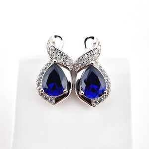 May include: A pair of silver earrings with a teardrop-shaped blue gemstone surrounded by smaller clear gemstones. The earrings have a twisted design.