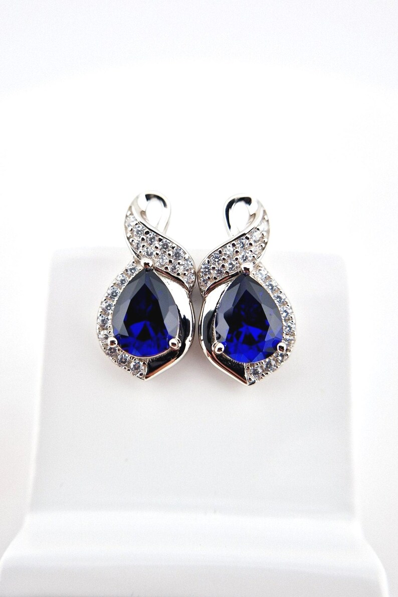 May include: A pair of silver earrings with a teardrop-shaped blue gemstone surrounded by smaller clear gemstones. The earrings have a twisted design.