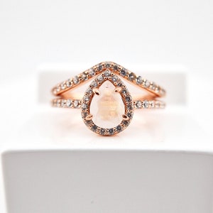 May include: A rose gold ring set with a pear-shaped gemstone surrounded by smaller stones. The ring has a delicate, v-shaped band.