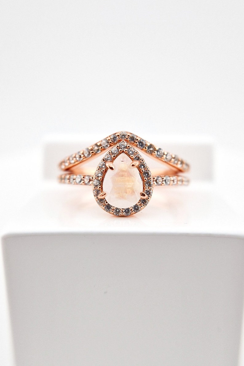 May include: A rose gold ring set with a pear-shaped gemstone surrounded by smaller stones. The ring has a delicate, v-shaped band.