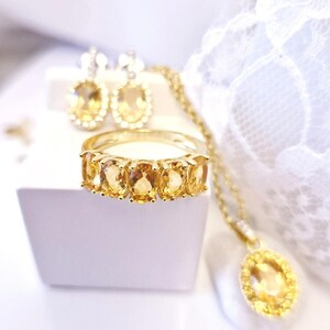 K&ouml;nnte beinhalten: Eine Sammlung goldfarbener Schmuckst&uuml;cke mit Citrin-Edelsteinen. Das Set umfasst einen Ring mit f&uuml;nf oval geschliffenen Steinen, passende Ohrringe und eine Anh&auml;ngerkette. Der Schmuck wird auf einer wei&szlig;en Oberfl&auml;che mit einem weichen, unscharfen Hintergrund pr&auml;sentiert.