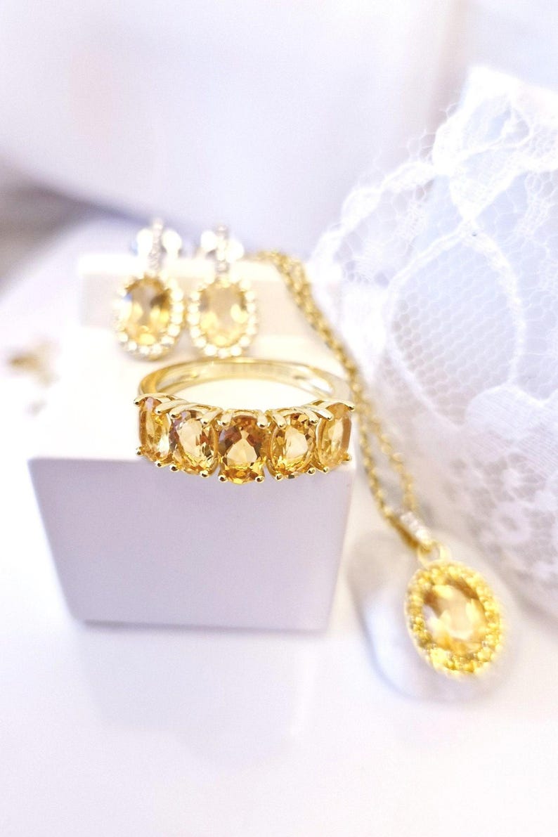 K&ouml;nnte beinhalten: Eine Sammlung goldfarbener Schmuckst&uuml;cke mit Citrin-Edelsteinen. Das Set umfasst einen Ring mit f&uuml;nf oval geschliffenen Steinen, passende Ohrringe und eine Anh&auml;ngerkette. Der Schmuck wird auf einer wei&szlig;en Oberfl&auml;che mit einem weichen, unscharfen Hintergrund pr&auml;sentiert.