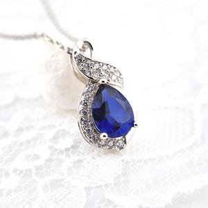 May include: A silver necklace with a pear-shaped blue gemstone surrounded by smaller clear gemstones. The pendant is in the shape of a swirl.