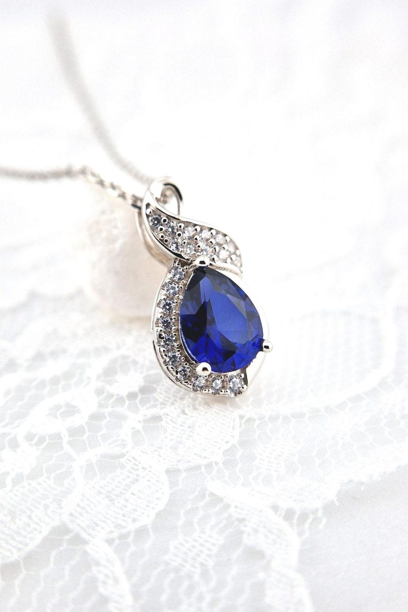 May include: A silver necklace with a pear-shaped blue gemstone surrounded by smaller clear gemstones. The pendant is in the shape of a swirl.