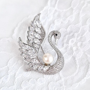 May include: A silver-toned swan brooch with a pearl and clear crystal accents. The swan's wings are detailed with marquise-cut crystals, and the body is encrusted with smaller crystals. The brooch is set against a white, lacy background.