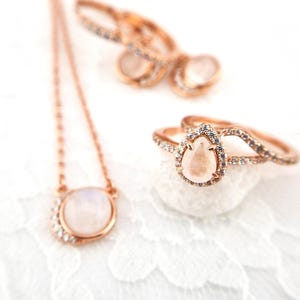 May include: A set of rose gold jewelry featuring moonstone gemstones. The set includes a necklace, a ring, and a pair of earrings. The jewelry is delicate and feminine, with a simple and elegant design.