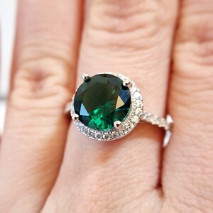 May include: A silver ring with a large, round, emerald green gemstone surrounded by a halo of smaller, clear gemstones.