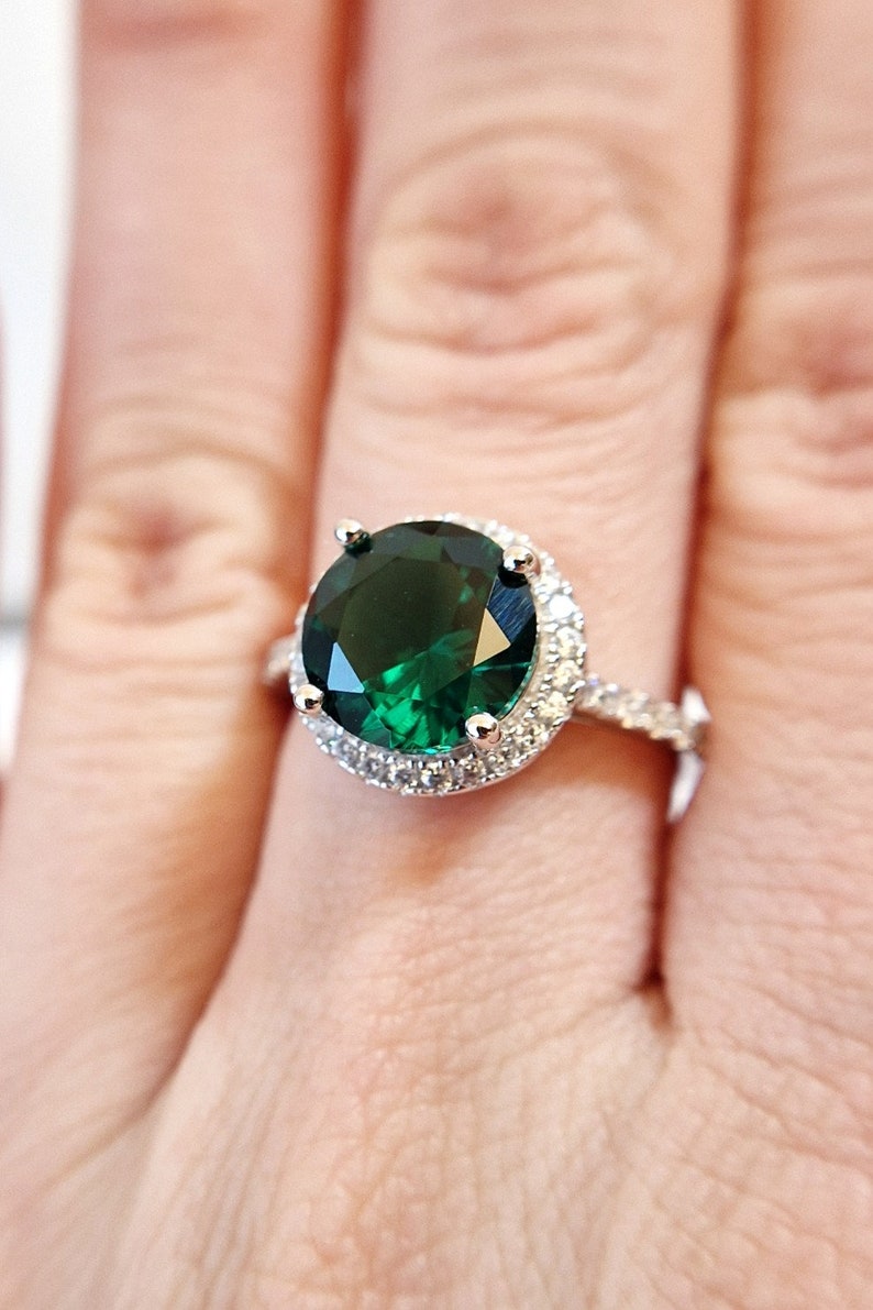May include: A silver ring with a large, round, emerald green gemstone surrounded by a halo of smaller, clear gemstones.