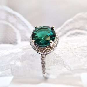 May include: A silver ring with a large, emerald green gemstone surrounded by a halo of smaller, clear gemstones.