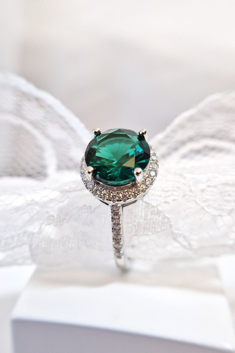May include: A silver ring with a large, emerald green gemstone surrounded by a halo of smaller, clear gemstones.