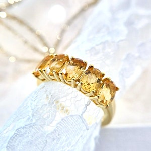 K&ouml;nnte beinhalten: Ein goldener Ring mit f&uuml;nf ovalen, goldgelben Edelsteinen. Der Ring wird auf wei&szlig;em Spitzenstoff mit verschwommenem Hintergrund pr&auml;sentiert. Die Edelsteine sind gleichm&auml;&szlig;ig auf einem goldenen Band angeordnet. Schmuck.
