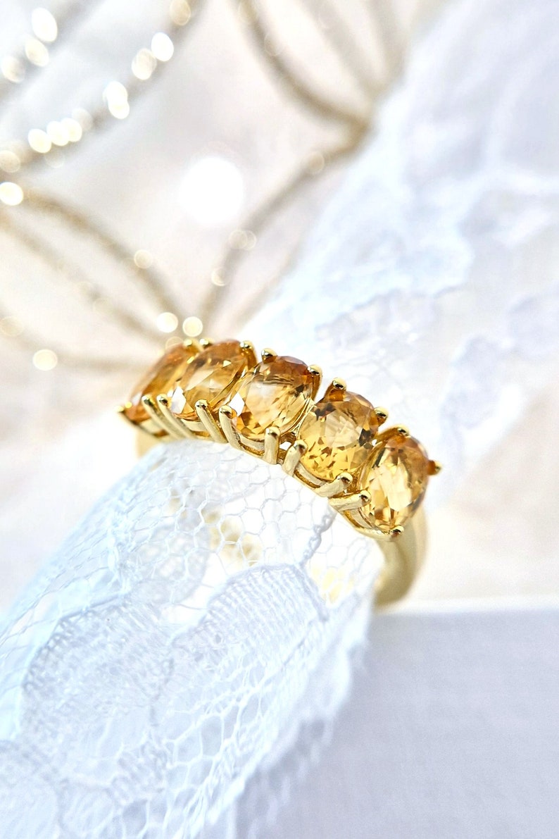K&ouml;nnte beinhalten: Ein goldener Ring mit f&uuml;nf ovalen, goldgelben Edelsteinen. Der Ring wird auf wei&szlig;em Spitzenstoff mit verschwommenem Hintergrund pr&auml;sentiert. Die Edelsteine sind gleichm&auml;&szlig;ig auf einem goldenen Band angeordnet. Schmuck.