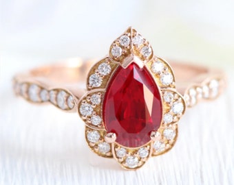 May include: A pear-shaped ruby gemstone ring set in a rose gold band. The ruby is surrounded by a halo of smaller diamonds and the band is also adorned with diamonds.