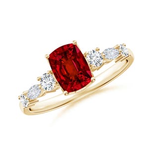 May include: A yellow gold ring featuring a central cushion-cut red gemstone held by four prongs. The band is accented on each side by a cluster of three clear, faceted stones, alternating between round and marquise shapes, set in yellow gold.