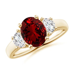 May include: A gold ring featuring a large, oval-cut, deep red gemstone flanked by two smaller, clear, oval-cut gemstones. The ring has a classic design with a polished gold band and prong settings.