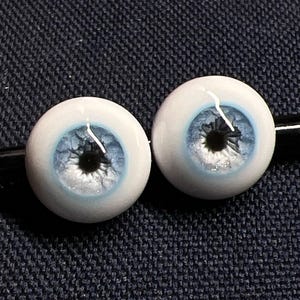 May include: Two doll eyes with a light blue iris and a black pupil. The eyes are round and white with a glossy finish. The eyes are set against a dark blue background.