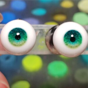 May include: Two green and blue doll eyes with a black pupil. The eyes are on a white background.