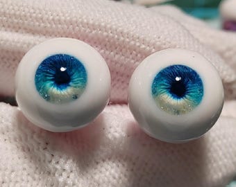 Puede incluir: Un par de ojos de muñeca azules y blancos con un iris blanco y una pupila azul. Los ojos están sobre una superficie blanca.