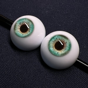 May include: A pair of white, spherical doll eyes featuring teal irises and dark brown pupils. The irises display a detailed, textured pattern with touches of gold and black. The eyes are set against a dark blue backdrop.