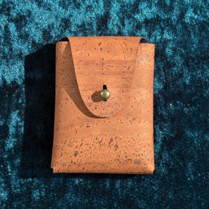 May include: A rectangular cork wallet with a flap closure and a small, round, green clasp. The wallet is a warm brown colour with darker speckles. The background is a dark teal colour.