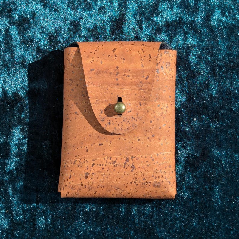 May include: A rectangular cork wallet with a flap closure and a small, round, green clasp. The wallet is a warm brown colour with darker speckles. The background is a dark teal colour.