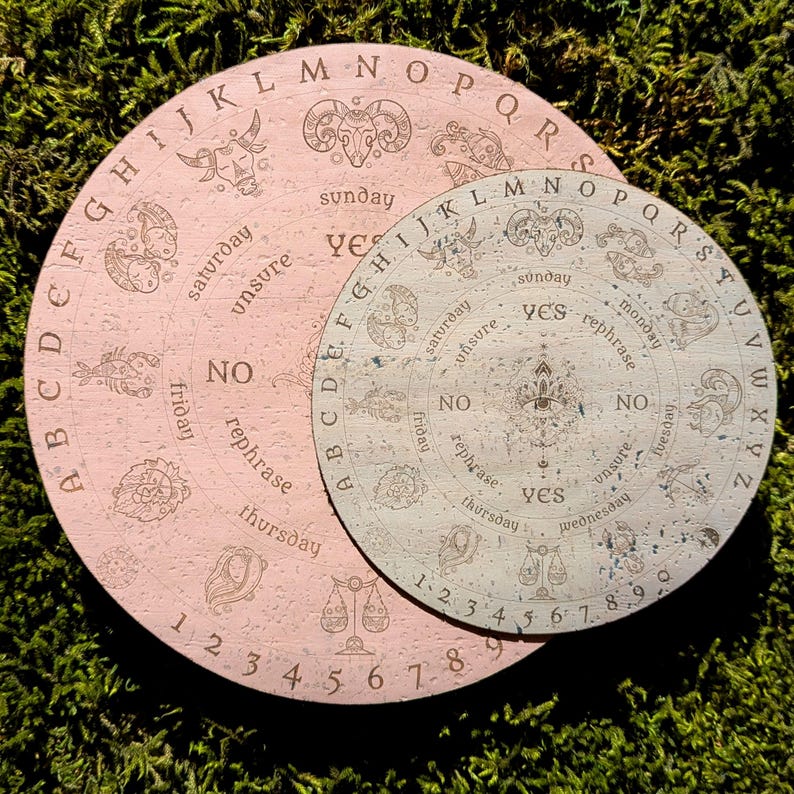 May include: Two circular divination boards. The larger board is pink with engraved letters, numbers, and zodiac symbols. The smaller board is a lighter color with similar engravings. Both have "yes", "no", and "unsure" options.