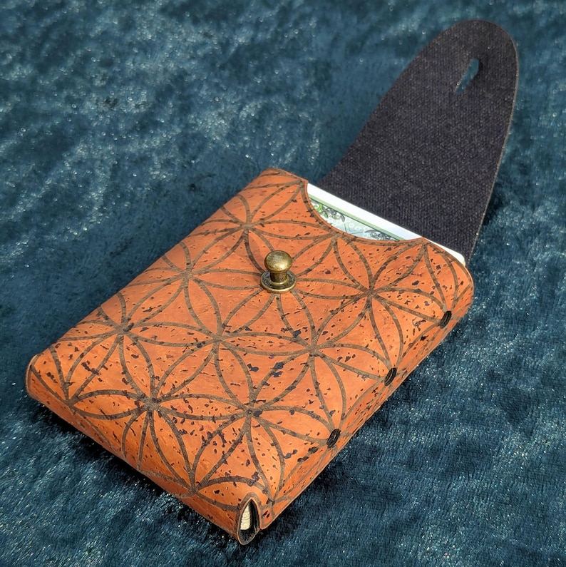 May include: A brown cork wallet featuring a flower of life pattern. The wallet has a brass clasp and a black interior. The wallet is open, revealing a glimpse of the contents inside.