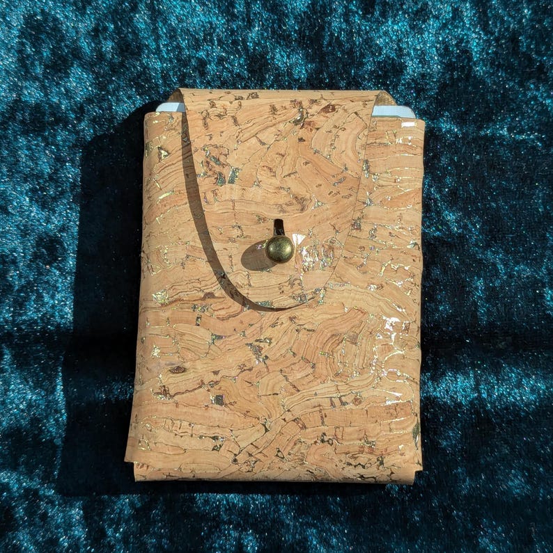 May include: A rectangular cork wallet with a flap closure and a brass-coloured button. The wallet has a natural cork pattern with gold flecks. The background is a dark teal fabric.